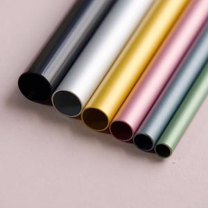 May include: An assortment of colorful, metallic tubes in varying sizes. The tubes are arranged diagonally, showcasing a palette of colors including black, silver, gold, pink, blue-grey, and green. The tubes are cylindrical with open ends, resting on a light pink surface.