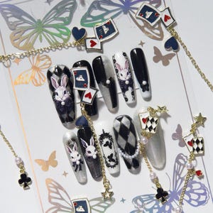 Könnte beinhalten: Ein Set aus künstlichen Fingernägeln mit schwarz-weiß-silbernen Designs, Hasenillustrationen, Spielkartenmotiven und Schmetterlingsakzenten. Das Set enthält goldfarbene Metallketten mit kleinen Stern-, Herz- und Kreuzanhängern an den Nägeln.