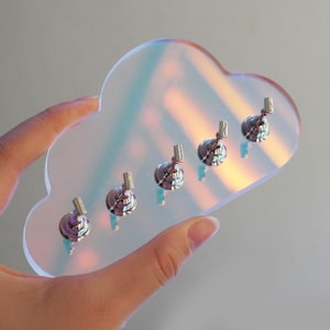 以下が含まれることがあります： 虹色の雲型キーホルダー。5つの銀色のフックが付いています。キーホルダーは、明るい灰色の背景に対して手で持たれています。雲は透明で反射性の高い素材で作られています。鍵を整理するのに最適です。
