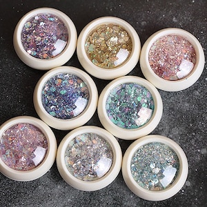 May include: Eight round containers with cream-colored rims, each filled with a unique blend of iridescent, metallic, and holographic nail art glitter flakes in various colors including purple, gold, pink, blue, green, and silver tones.