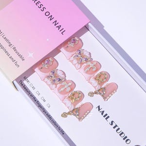 May include: A set of press-on nails in a box. The nails are light pink with white floral accents and various rhinestone embellishments, including butterflies and crosses. The box is pink and white with the text "PRESS ON NAIL" and "NAIL STUDIO".