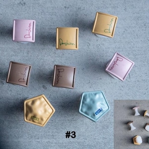 May include: A collection of decorative cabinet knobs in different shapes and shades. Square knobs display the word "Fashion", while others are pentagonal with the word "Love". Colours include gold, brown, and pink. A small group of additional knobs is also visible.