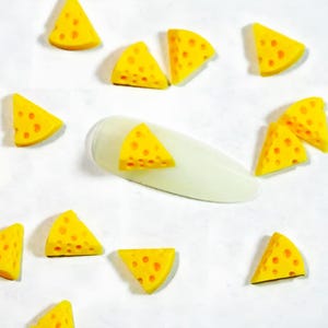 May include: A collection of small, triangular yellow cheese-shaped charms with scattered holes, arranged around a single, smooth, elongated, translucent white nail art display tip featuring one of the cheese charms attached to its surface.