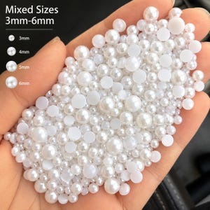 Puede incluir: Una colección de perlas artificiales blancas, redondas y de base plana en tamaños mixtos que van de 3 mm a 6 mm. El texto en la imagen indica Mixed Sizes 3mm-6mm, con guías visuales para los tamaños de 3 mm, 4 mm, 5 mm y 6 mm. Son cuentas blancas.