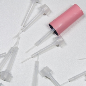 Puede incluir: Una colección de puntas aplicadoras de plástico transparente con una tapa rosa. Las puntas tienen un diseño largo y delgado, y la tapa es cilíndrica. Los artículos están dispersos sobre una superficie blanca.