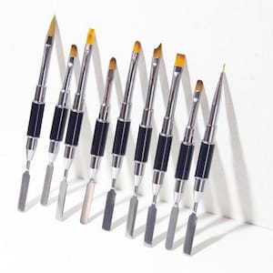 May include: An assortment of nail art tools, including double-ended brushes and spatulas. The tools have silver metal handles with black grips and yellow brush tips. The tools are arranged on a white surface.