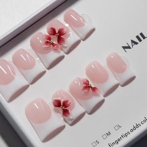 May include: A set of artificial nails with a French manicure design. The nails are white with a pink oval shape and adorned with red flower accents. The box reads "NAIL STUDIO" and "fingertips adds colors to life".