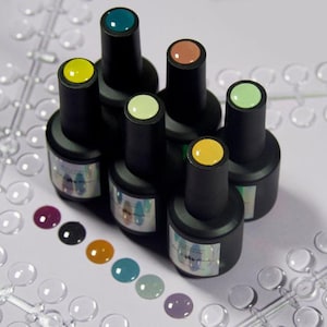 May include: A collection of gel nail polish bottles in various colors, including yellow, teal, and peach. The bottles are black with colored tops and are arranged on a white surface. A color swatch of nail polish is displayed below the bottles.