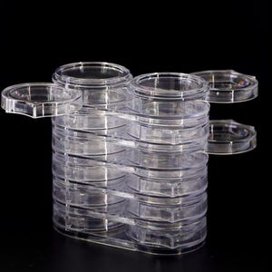 May include: A stack of clear, transparent plastic coin holders. Each holder has a circular design with a lid, and they are arranged in a vertical stack. The holders are designed to protect and display coins.