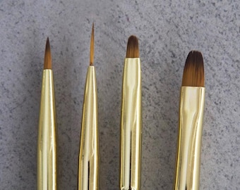 May include: Four gold-colored paint brushes with brown bristles. The brushes have different tip shapes, including a fine point, a thin, angled tip, and two flat, rounded tips. The handles are smooth and shiny, reflecting the light.
