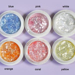 May include: Six small round white containers filled with iridescent, flaky nail art decorations. The colors are labeled as blue, pink, white, orange, coral, and yellow. Each container holds a collection of thin, shimmering, reflective flakes.