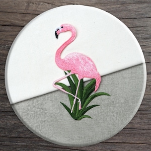 May include: Pink flamingo embroidered on a white and grey fabric background. The flamingo is standing on a green plant.