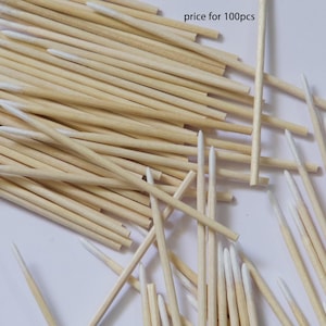 May include: A collection of wooden toothpicks with white tips, scattered on a light surface. The toothpicks are made of natural wood and are pointed at one end. The image shows a close-up view of the toothpicks.