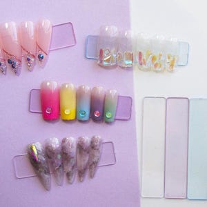 May include: A collection of artificial nails in various designs and colors. Some nails are clear with iridescent accents, others feature gradient colors, and some have decorative gems. The nails are displayed on clear plastic holders.