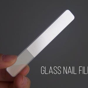 Puede incluir: Una lima de uñas de vidrio transparente con una superficie de limado texturizada. La lima es rectangular con bordes redondeados y se sostiene sobre un fondo gris. El texto "GLASS NAIL FILE" se muestra en blanco.