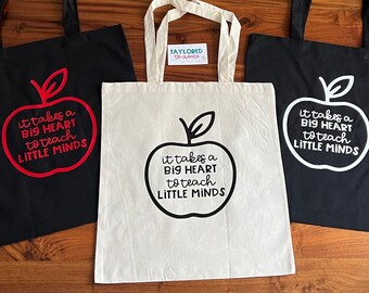 Può includere: Tre borse a tracolla con la scritta "ci vuole un grande cuore per insegnare alle piccole menti" stampata su di esse. Le borse sono nere con disegni di mele rosse e bianche. Una borsa è bianca con un disegno di mela nera.