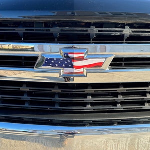 May include: Close-up of a black vehicle grille featuring a chrome Chevrolet emblem. The emblem is filled with a design of the American flag, showcasing the stars and stripes. The grille has horizontal black slats.