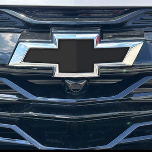 May include: Close-up of a black car grille featuring the Chevrolet logo in chrome. The logo is a black bow tie shape with a chrome border. A small camera is centered below the logo.