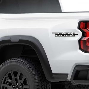 May include: A white pickup truck with a black "Trail Boss" decal on the side of the bed. The truck has black wheels and a black bumper.