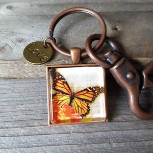 Puede incluir: Un llavero de cobre con un encanto de vidrio cuadrado que presenta una mariposa monarca sobre una flor. El encanto est&aacute; unido a un anillo de cobre y un cierre de langosta. Un peque&ntilde;o encanto de cobre con la palabra "Love" tambi&eacute;n est&aacute; unido al anillo.