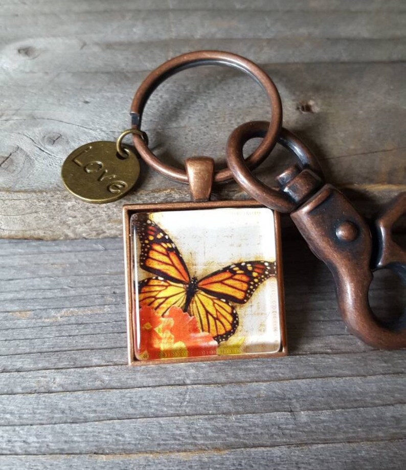 Puede incluir: Un llavero de cobre con un encanto de vidrio cuadrado que presenta una mariposa monarca sobre una flor. El encanto est&aacute; unido a un anillo de cobre y un cierre de langosta. Un peque&ntilde;o encanto de cobre con la palabra "Love" tambi&eacute;n est&aacute; unido al anillo.