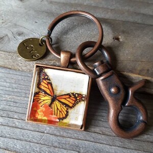 Puede incluir: Un llavero de tono cobre con un encanto de vidrio cuadrado que presenta una mariposa monarca sobre una flor. El encanto est&aacute; unido a un anillo dividido con un encanto m&aacute;s peque&ntilde;o que dice "Love".