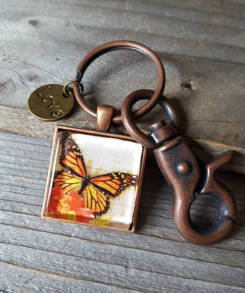 Puede incluir: Un llavero de tono cobre con un encanto de vidrio cuadrado que presenta una mariposa monarca sobre una flor. El encanto est&aacute; unido a un anillo dividido con un encanto m&aacute;s peque&ntilde;o que dice "Love".