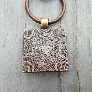Puede incluir: Un llavero de tono cobre con un colgante cuadrado que presenta un dise&ntilde;o circular de c&iacute;rculos conc&eacute;ntricos. El colgante est&aacute; unido a un anillo partido y un broche de langosta.