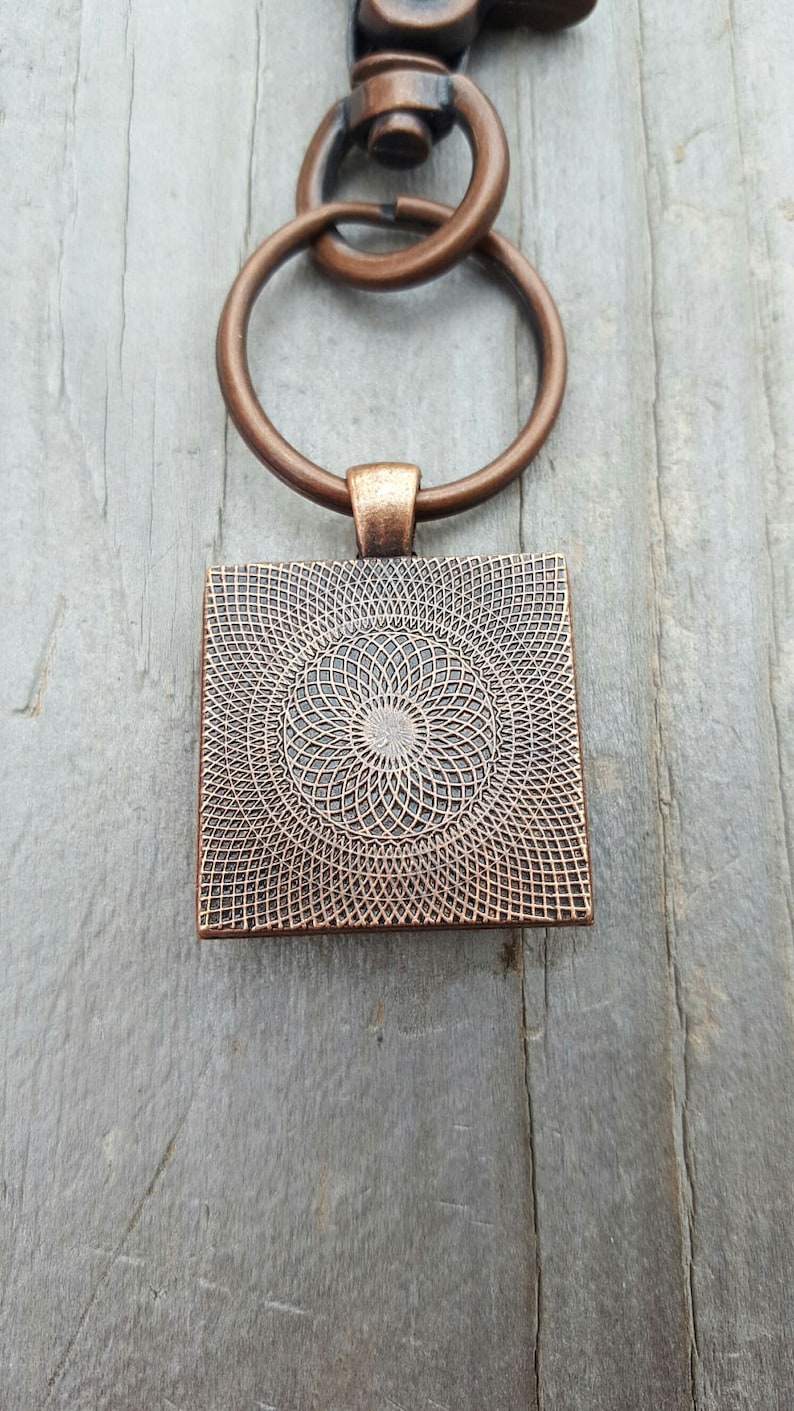 Puede incluir: Un llavero de tono cobre con un colgante cuadrado que presenta un dise&ntilde;o circular de c&iacute;rculos conc&eacute;ntricos. El colgante est&aacute; unido a un anillo partido y un broche de langosta.