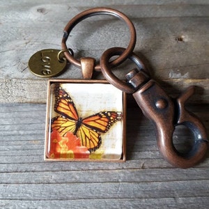 Puede incluir: Un llavero de tono cobre con un dije cuadrado que presenta una mariposa monarca sobre un fondo floral. El dije est&aacute; unido a un anillo dividido y un broche de langosta. El llavero tambi&eacute;n incluye un peque&ntilde;o dije redondo con la palabra "Love" grabada en &eacute;l.