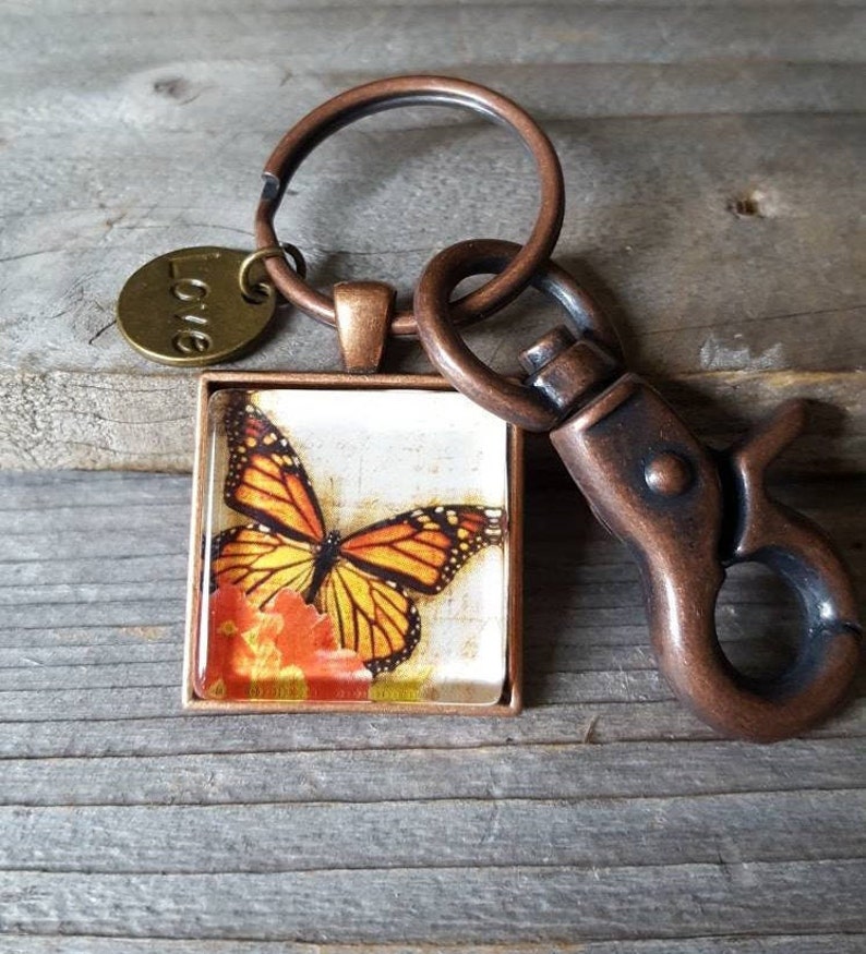 Puede incluir: Un llavero de tono cobre con un dije cuadrado que presenta una mariposa monarca sobre un fondo floral. El dije est&aacute; unido a un anillo dividido y un broche de langosta. El llavero tambi&eacute;n incluye un peque&ntilde;o dije redondo con la palabra "Love" grabada en &eacute;l.