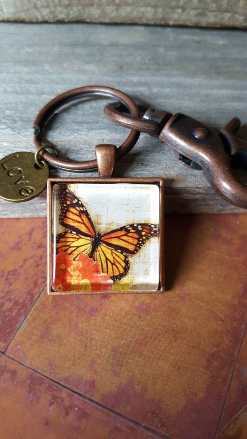 Puede incluir: Un llavero de cobre con un encanto de vidrio cuadrado que presenta una mariposa monarca sobre un fondo floral. El encanto est&aacute; unido a un anillo de cobre con un peque&ntilde;o encanto redondo que dice "Love".
