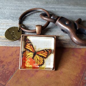 Puede incluir: Un llavero de tono cobre con un dije de vidrio que presenta una mariposa monarca sobre un fondo floral. El dije est&aacute; unido a un anillo dividido y un broche de langosta. Una peque&ntilde;a etiqueta de tono cobre con la palabra "Love" est&aacute; unida al anillo dividido.