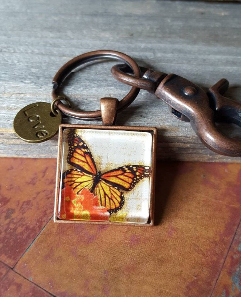Puede incluir: Un llavero de tono cobre con un dije de vidrio que presenta una mariposa monarca sobre un fondo floral. El dije est&aacute; unido a un anillo dividido y un broche de langosta. Una peque&ntilde;a etiqueta de tono cobre con la palabra "Love" est&aacute; unida al anillo dividido.