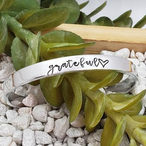 May include: A silver cuff bracelet with the word "grateful" and a small heart engraved on it. The bracelet is lying on a bed of small white rocks.