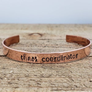 May include: A copper cuff bracelet with the words "chaos coordinator" stamped across the front. The bracelet is a warm, reddish-brown color and sits on a wooden surface.
