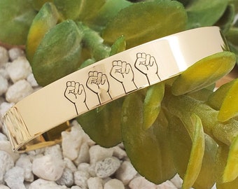 May include: Gold-toned metal bracelet with five raised fists engraved on the surface.
