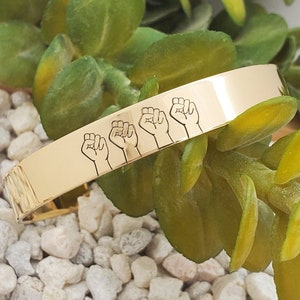 May include: Gold-toned metal bracelet with five raised fists engraved on the surface.