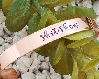 May include: Rose gold bangle bracelet with the words "shit show" engraved on it.