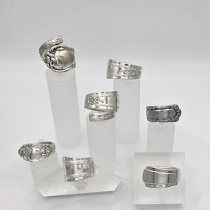May include: A collection of handcrafted silver rings, each uniquely designed from repurposed silverware. The rings showcase intricate floral and ornate patterns, displayed on clear acrylic stands against a white backdrop. The rings are a unique jewelry item.