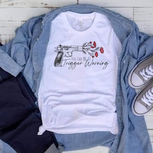 May include: White t-shirt with a black and white illustration of a gun with red flowers and the text "You Had Me At Trigger Warning".