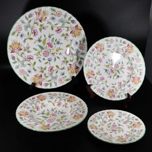 May include: A set of four white ceramic plates with a floral pattern in pink, yellow, and blue. Each plate has a green rim. The plates are displayed on a black surface.
