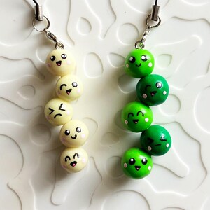 May include: Two beaded phone charms. One charm features a string of off-white beads with painted faces. The other charm has green beads with painted faces. Both charms have black cords and silver-colored metal attachments.