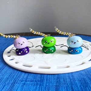 May include: Three stacked, round, clay charms with cute faces. The charms are purple, green, and blue. Each charm has a gold chain attached.