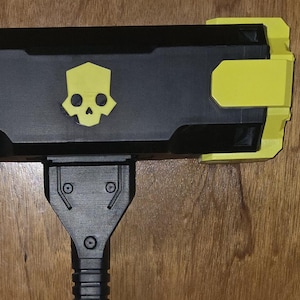 May include: A black sledgehammer with a long, textured handle and a rectangular head. The head features a yellow skull icon on the front and a bright yellow accent piece on the right side. The handle has a ribbed grip section and a textured base.