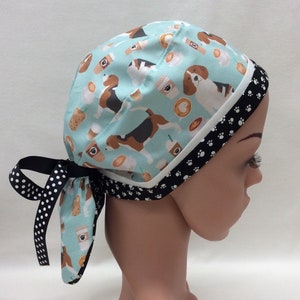 May include: A blue and white patterned surgical scrub hat with a black and white polka dot tie. The hat features a repeating pattern of beagles and coffee cups. The hat is worn on a mannequin head.