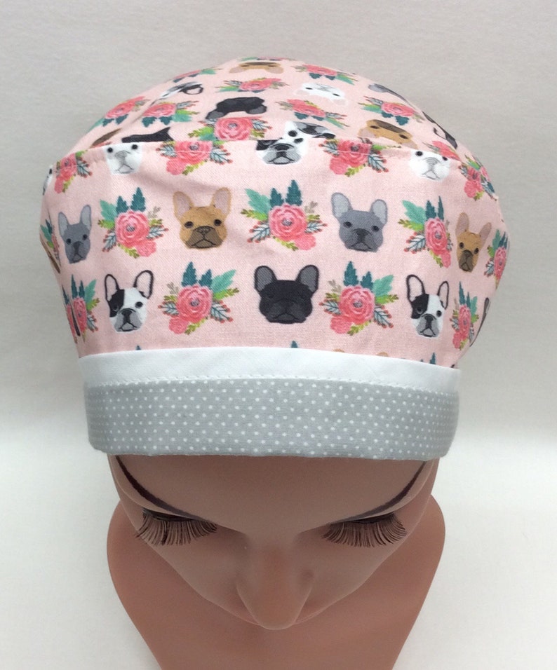 May include: A pink scrub hat with a white band and gray polka dot trim. The hat features a repeating pattern of French Bulldogs and pink flowers.