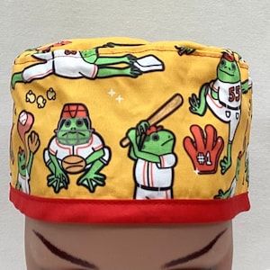 May include: Yellow scrub hat with a red trim featuring a repeating pattern of green frogs playing baseball. The frogs are wearing baseball uniforms with the number 55 on one frog. The hat has a red trim around the bottom.