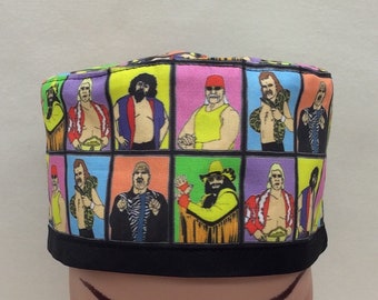 May include: A colorful, square-shaped fabric hat featuring a grid of cartoon illustrations of wrestlers. The hat has a black brim and a variety of bright background colors, including yellow, pink, and purple. The wrestlers are depicted in various poses.