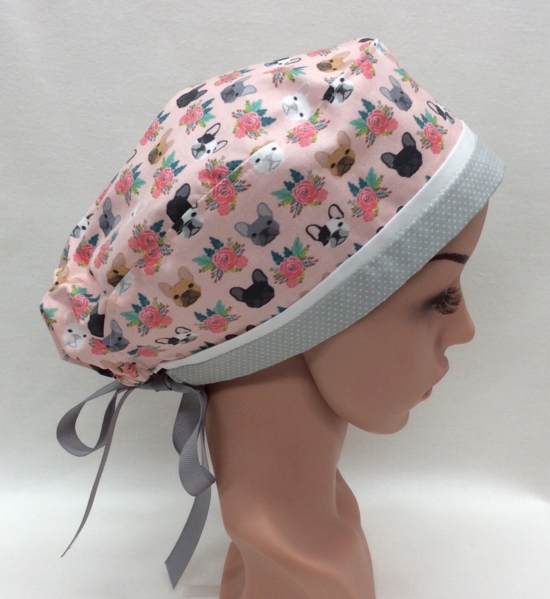 May include: A pink surgical scrub hat with a white and gray trim. The hat has a pattern of French bulldog faces and pink flowers. The hat has a gray ribbon tie.